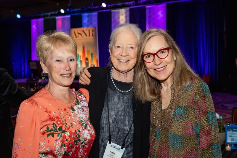 Three women smiling at a social event with an ASNR sign in the background.