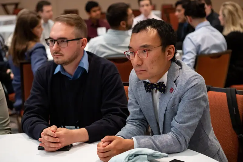 Two men sitting at a table attentively listening to an unseen speaker.