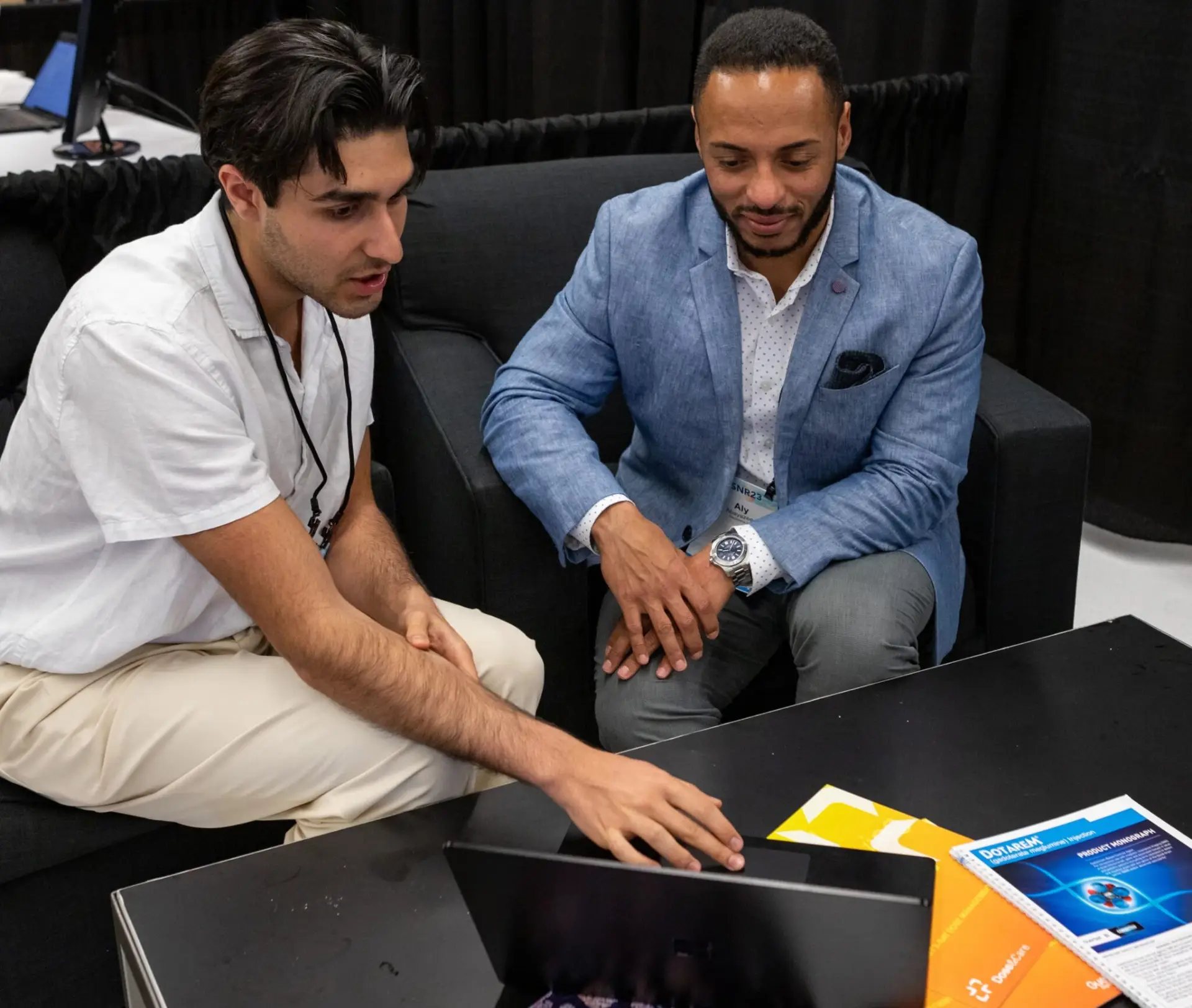 Two men sitting on a black couch, discussing something on a laptop. Various colorful booklets are on the coffee table in front of them.